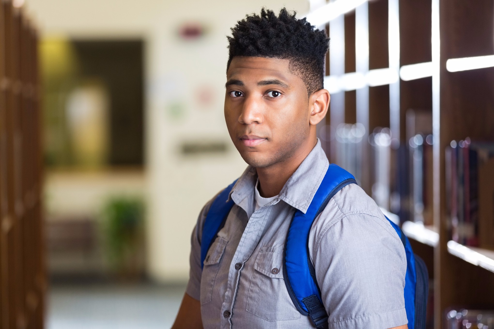 Teenage African American high school boy is looking at the camera with a serious expression. Student is standing in between rows of bookshelves in school library. He is wearing a backpack and casual shirt.